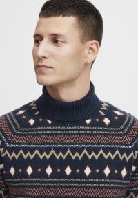 Pulover navy cu guler înalt, prezentând modele geometrice în crem, portocaliu și maro. Textură din fleece, cu guler și umeri cu nervuri.