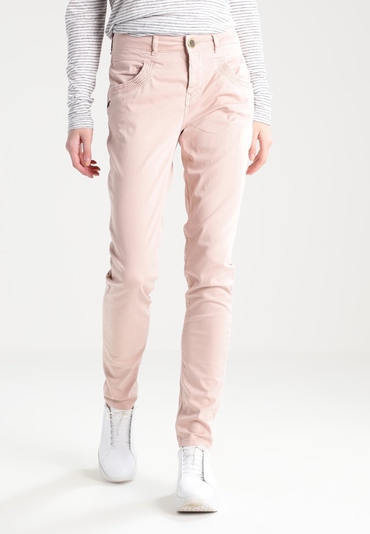 Jasnoróżowe, dopasowane spodnie o gładkiej teksturze; posiadają przednie kieszenie z delikatnym przeszyciem; zestawione z białymi, niskimi trampkami i paskowanym topem.