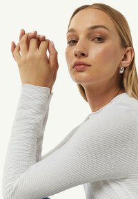 Biała bluzka z długim rękawem o fakturowanej powierzchni z okrągłym dekoltem. Modelka pozuje z dłońmi złożonymi, nosząc srebrne kolczyki wkrętki. Naturalny makijaż i proste włosy.