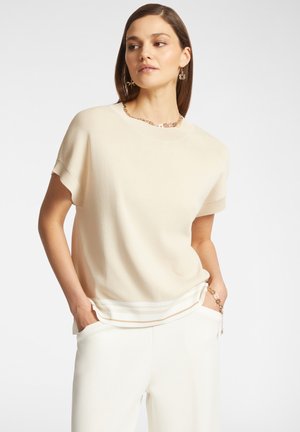 Maglione beige a maniche corte con scollatura rotonda e orlo a coste, indossato con pantaloni bianchi. Texture liscia, design minimalista, vestibilità casual.