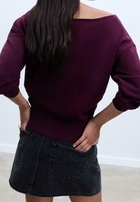 Pull bordeaux à épaules dénudées avec manches longues, associé à une mini-jupe en denim noir dotée de deux poches arrière et de détails de couture subtils.