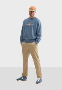 Sudadera azul con texto impreso, pantalones caqui y zapatillas azules con cordones blancos. Gorra naranja con texto. Tela de algodón, estilo casual.