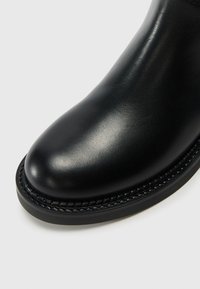 Bota de cuero negro con punta redondeada, textura suave y costuras detalladas alrededor de la suela. Muestra de artesanía y durabilidad.