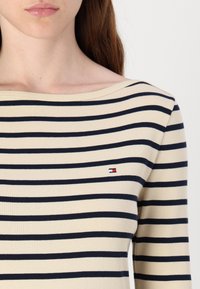 Pruhovaný svetřík s dlouhým rukávem v krémové a námořnické modři, s lodičkovým výstřihem a malým logem v rohu. Měkká pletená textura a přiléhavý střih.
