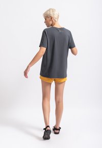 Grijze katoenen t-shirt met korte mouwen en een ronde hals. Heeft een klein logo op de bovenrug. Draagt bij gele shorts en sandalen.