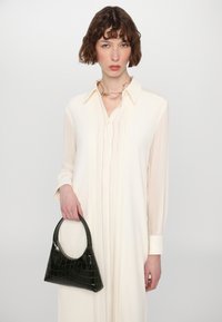 Bolso de mano verde de imitación de cocodrilo con un asa curva, sostenido por una persona que lleva un vestido camisero plisado color crema con mangas largas.