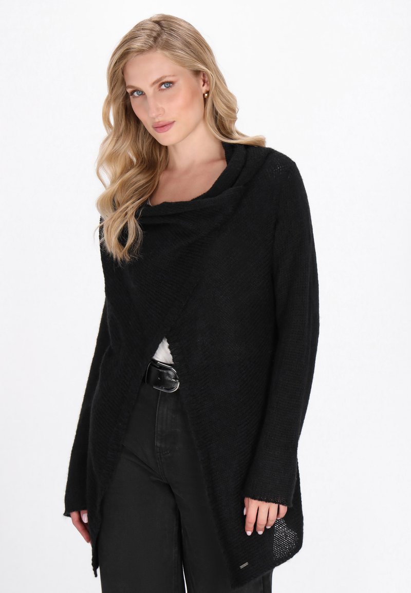 Blonde Frau in einer schwarzen strukturierten Wickeljacke über schwarzen Hosen mit einem schwarzen Gürtel, stehend vor einem weißen Hintergrund.