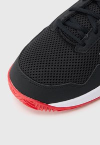 Zwarte sportschoen met een ademende mesh bovenkant, rode rubberen zool en platte zwarte veters met subtiele ontwerpdetails aan de zijkanten.