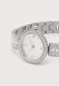 Ασημένιο ρολόι χειρός με στεφάνη και βραχιόλι διακοσμημένα με κρύσταλλα, λευκό καντράν με ελάχιστους δείκτες και μικρό κορώνα στο πλάι.