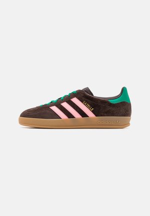 Zapatillas de ante marrón con tres rayas rosas, detalles en verde y suela de goma. Cuentan con cierre de cordones y el logo "Gazelle" en el lateral.