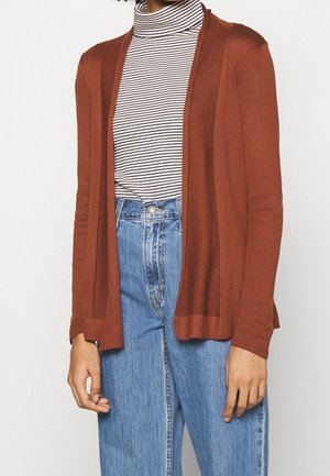 Persoon die lichtblauwe jeans met hoge taille, een zwart-wit gestreepte coltrui en een open roestkleurige cardigan draagt, staat tegen een witte achtergrond.