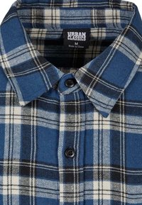 Flanelska košulja s plavim, crnim i bijelim kockastim uzorkom. Ima kragnu na dugmad i crne detalje na dugmadima. Etiketa kaže "Urban Classics", veličina M.
