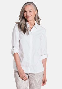 Weißes Hemd mit gekrempelten Ärmeln, glatte Textur, klassischer Kragen. Kombiniert mit gemusterten beigen Hosen mit horizontalen Linien.