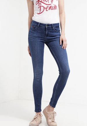 Femme portant un jean skinny bleu foncé, un t-shirt blanc à motif graphique et des baskets beiges, debout devant un fond blanc uni.