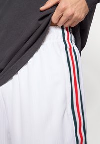 Λευκό αθλητικό σορτς με κάθετη ανάγλυφη υφή, διακοσμημένο με κόκκινες, πράσινες και λευκές ρίγες κατά μήκος των πλευρών. Γκρι πουκάμισο ορατό εν μέρει.