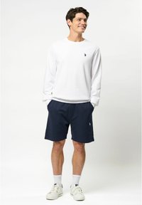 Witte longsleeve shirt met een klein zwart logo, gecombineerd met marineblauwe shorts met een elastische tailleband en zijzakken. Witte sneakers.