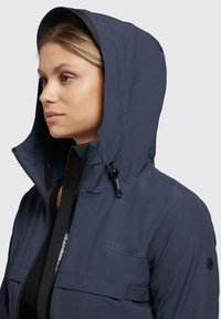 Navyblauwe capuchonjack met een rits aan de voorkant, verstelbare trekkoordcapuchon en een strakke, lichtgewicht ontwerp. Bevat een logo op de borst en zakdetails.