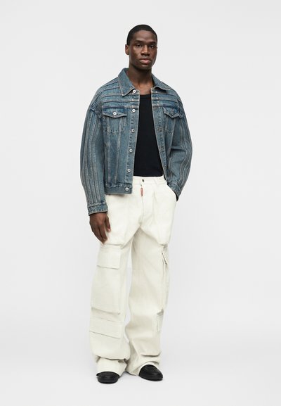 Άνδρας που στέκεται φορώντας ριγέ τζιν μπουφάν, μαύρο πουκάμισο, λευκό παντελόνι cargo με μεγάλες τσέπες και μαύρα παπούτσια, πάνω σε απλό λευκό φόντο.