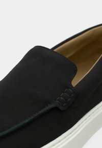 Μαύρο παπούτσι τύπου slip-on με λεία, ματ επιφάνεια και αντίθετη λευκή σόλα· διαθέτει διακοσμητική ραφή κατά μήκος της άνω ακμής και μαλακό εσωτερικό.