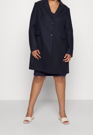 Woman wearing a knee-length dark navy coat over a dress with beige open-toe block heel sandals, standing against a plain white background.
