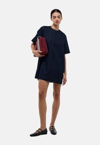 Robe chemise en denim bleu foncé, à manches courtes, décolleté rond, avec une coupe droite. Le mannequin tient un sac bordeaux avec des rayures blanches. Chaussures noires.