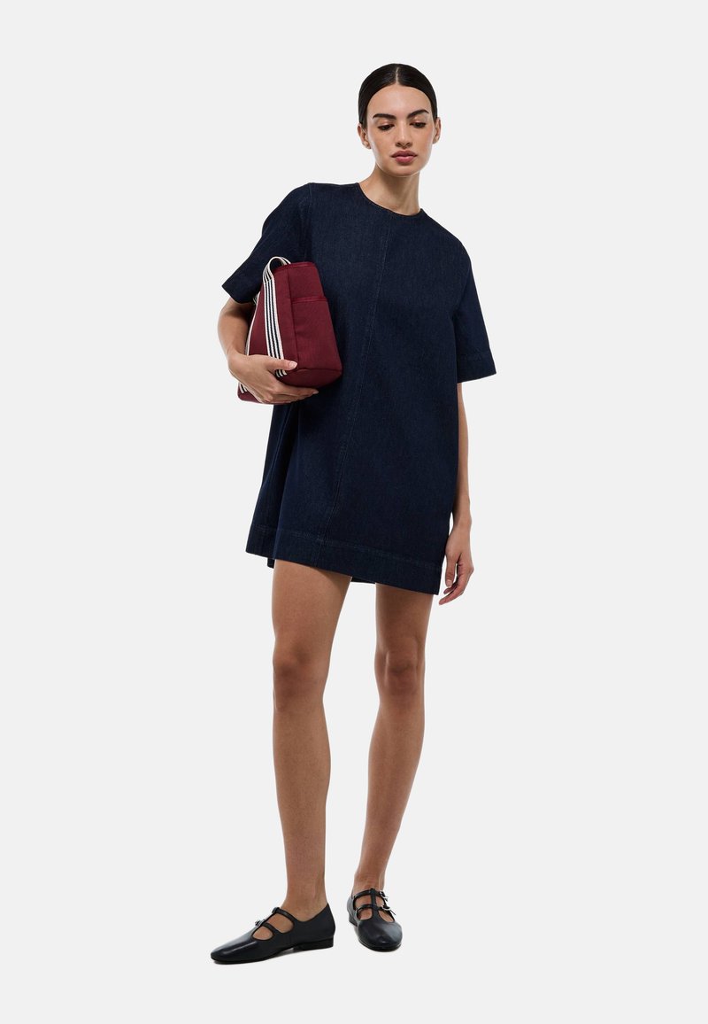 Robe chemise en denim bleu foncé, à manches courtes, décolleté rond, avec une coupe droite. Le mannequin tient un sac bordeaux avec des rayures blanches. Chaussures noires.