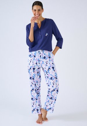 Lachende vrouw die blootsvoets staat, gekleed in een marineblauw shirt met lange mouwen en wijde broek met bloemenpatroon, met haar hand in de zak.