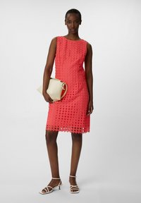 Korallfarbendes ärmelloses Kleid mit einem Diamantmuster, knielanger Saum, kombiniert mit einer cremefarbenen Handtasche und weißen Riemchenheels.