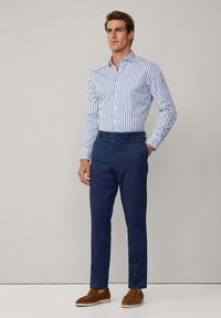 Camicia bianca con righe verticali blu, maniche lunghe, davanti con bottoni; abbinata a pantaloni blu navy e mocassini marroni. Texture liscia in tutto il capo.
