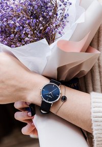 Montre à bracelet noir avec un cadran bleu foncé et des accents argentés, portée au poignet, à côté d'un délicat bracelet en argent. Fleurs séchées violettes regroupées.