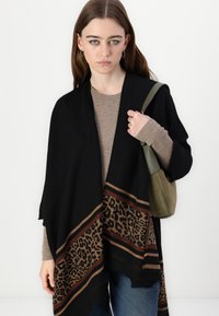 Sciarpa nera con bordo a stampa leopardata, caratterizzata da strisce marrone scuro e marrone chiaro; abbinata a un top beige a righe sottili e una borsa verde.