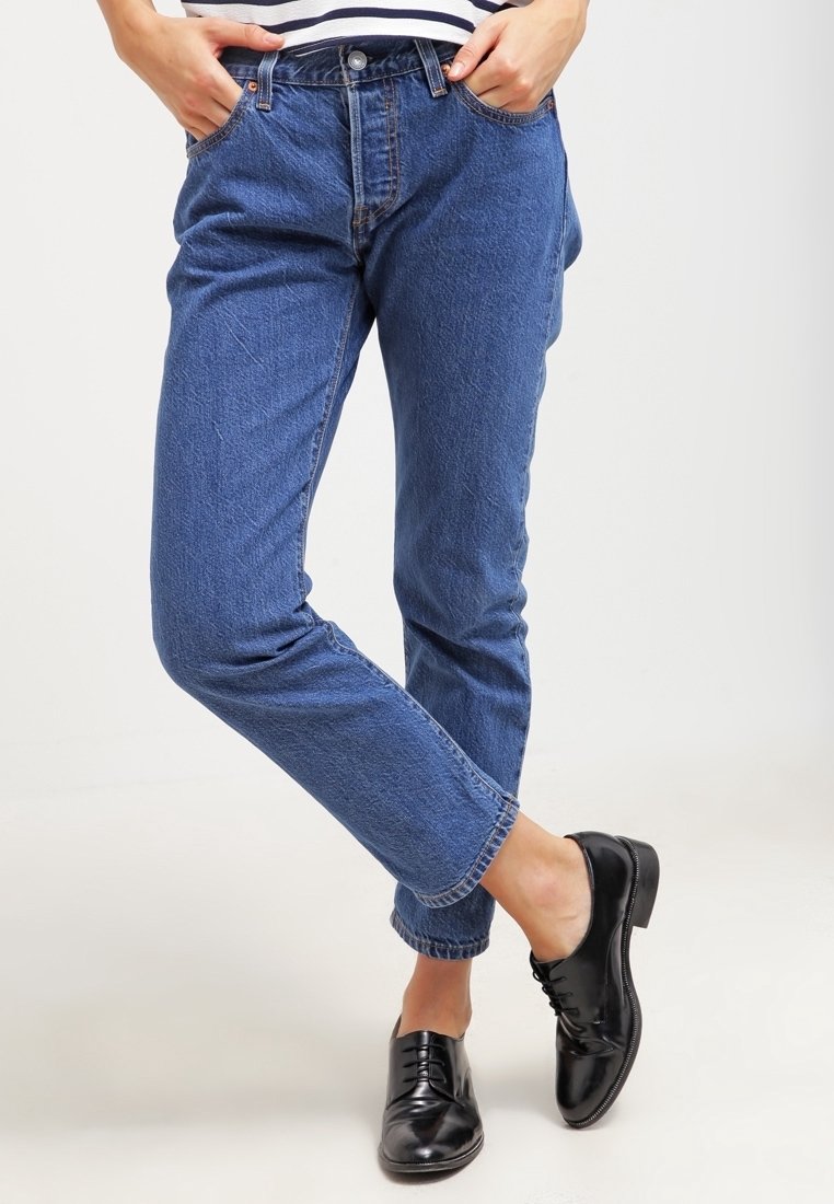 Jean en denim bleu avec une coupe ample, présentant un bouton et une fermeture éclair, cinq poches et des ourlets retroussés, associés à des chaussures noires à lacets.