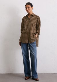 Chemise à carreaux marron et beige avec des manches longues, patte de boutonnage à l'avant et poches sur les côtés ; associée à un jean bleu à jambes larges et des chaussures marron.