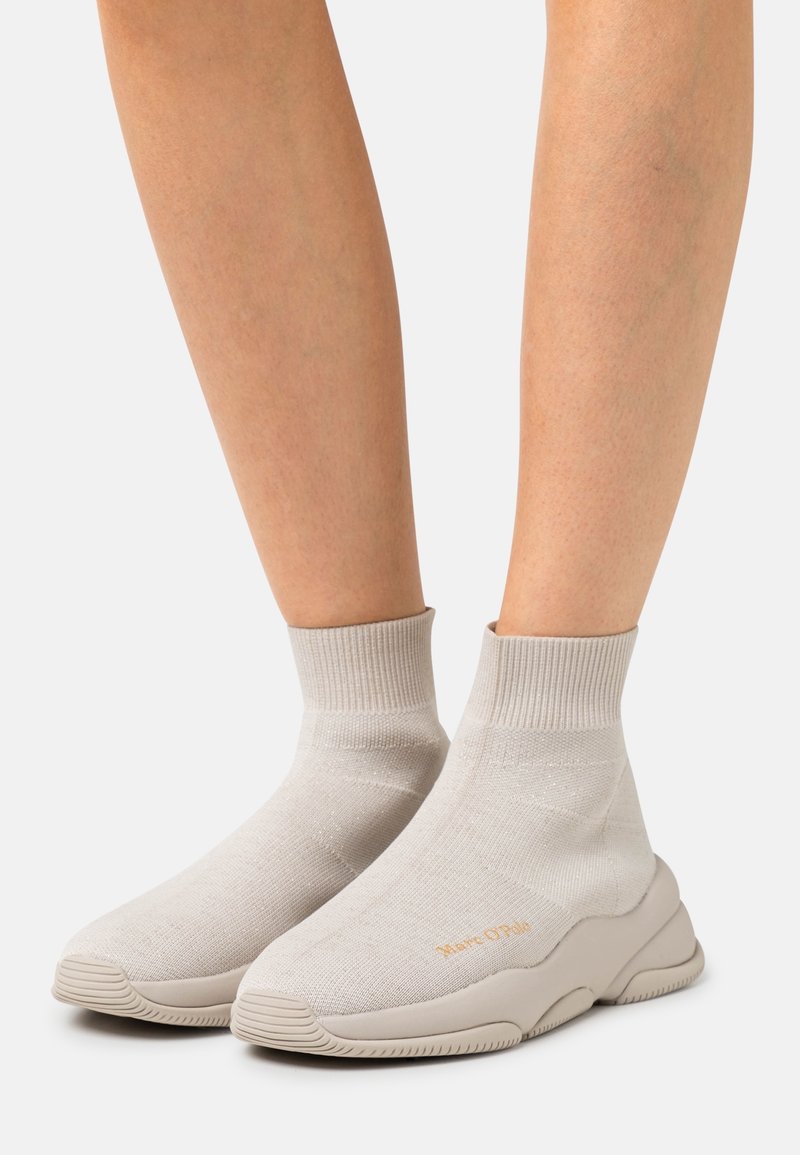 Baskets beige style chaussette avec bord-côtes à la cheville et semelles épaisses texturées portées sur jambes nues sur fond blanc.