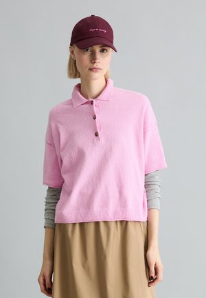 Jeune femme portant une casquette bordeaux et un polo rose à manches courtes superposé à un t-shirt gris à manches longues, avec une jupe beige, sur un fond gris uni.