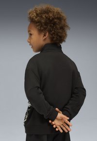 Bambino con capelli ricci che indossa una felpa nera, rivolto a sinistra, con le mani unite dietro la schiena, su uno sfondo grigio uniforme.