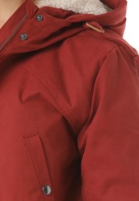 Veste rouge avec un extérieur texturé, une capuche doublée et des boutons-pression. Les caractéristiques incluent des détails matelassés et une doublure intérieure douillette.