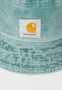 Teal kordfløjl bucket hat med et vævet mærke med et gult logo og "carhartt" tekst. Texturerede lodrette striber, blødt stof.