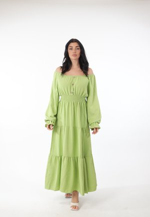 Kvinde går fremad iført en lang, lysegrøn off-shoulder kjole med lange ærmer og beige sandaler med åbne tæer.