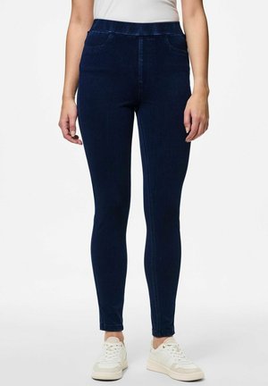 Donkerblauwe denim leggings met een gladde textuur en een elastische tailleband. Beschikt over twee voorzakken en een slim fit ontwerp. Gemaakt in combinatie met witte sneakers.