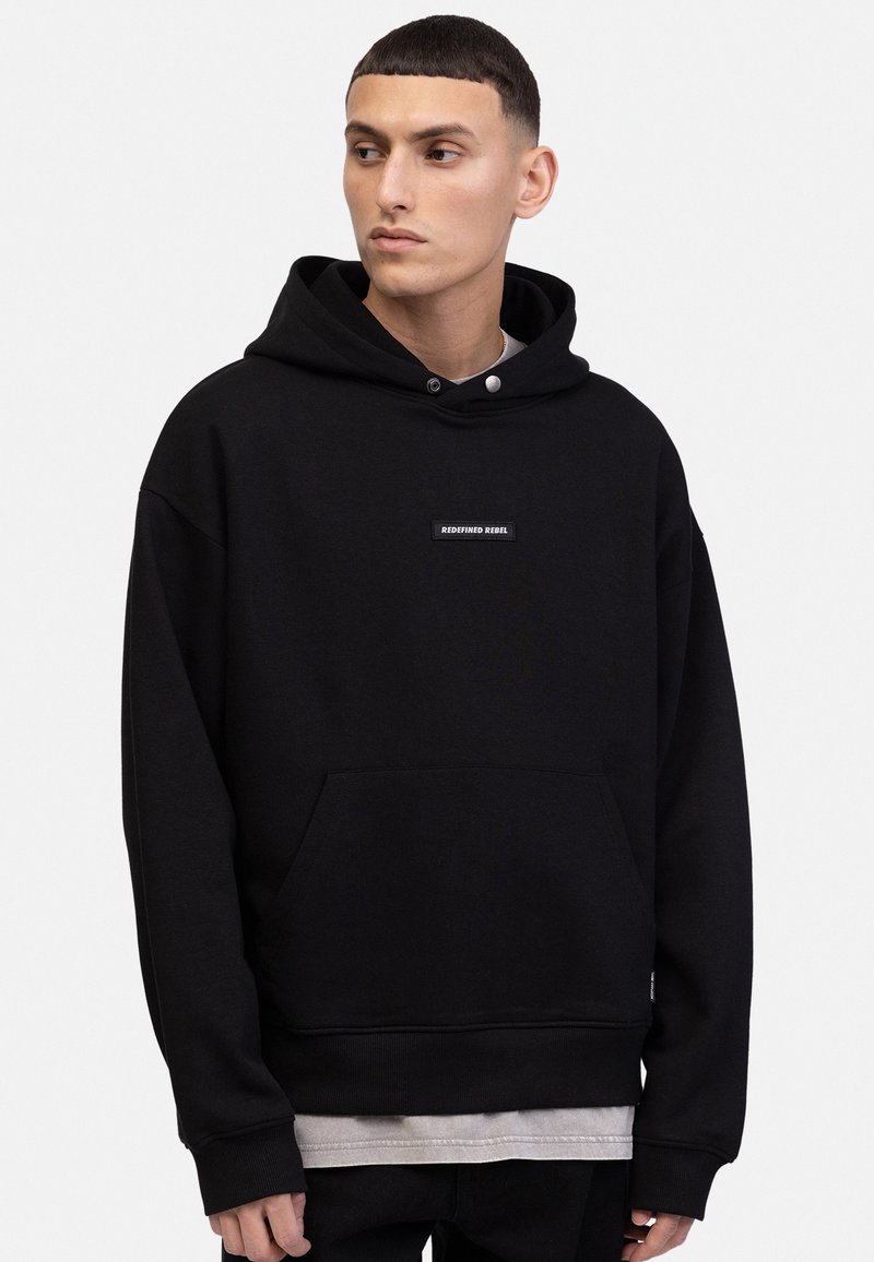 Haut de survêtement en polaire noire avec une poche avant, arborant une petite étiquette "REDEFINED REBEL". La capuche est dotée d'un cordon de serrage et d'une texture subtile.
