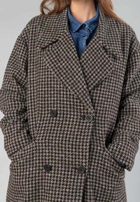 Abrigo de patrón houndstooth en tonos marrón y negro, con diseño de doble botonadura, grandes solapas y dos bolsillos frontales visibles. Detalles de botones.