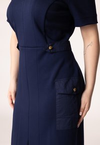 Dunkelblaues Kleid mit kurzen Ärmeln, das ein tailliertes Design, texturierten Stoff und seitliche Taschen mit goldenen Knopfakzenten aufweist.
