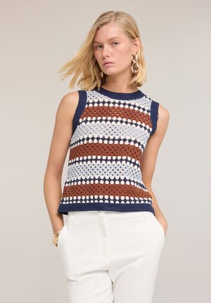 Blondynka w bezrękawnym, paskowanym topie na szydełku w kolorach granatu, brązu, bieli i jasnoniebieskiego, zestawiona z białymi spodniami i złotymi kolczykami-kołami.