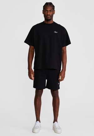 Mann steht frontal, trägt ein lockeres schwarzes T-Shirt, schwarze Shorts und weiße Turnschuhe vor einem einfarbigen hellen Hintergrund.