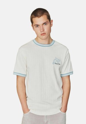 Young man with short hair wearing a white ribbed T-shirt with light blue striped collar and sleeves and a small fish logo on chest.