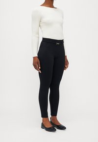 Svarta slim-fit byxor med en slät textur, med en guldfinish i midjan, kombinerat med en ribbad vit långärmad topp.