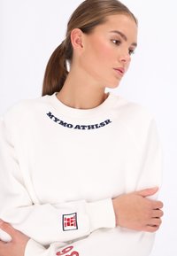 Weißer Pullover mit marineblauem, gesticktem Text am Halsausschnitt und rotem grafischem Detail am Ärmel. Weiche Textur und entspannte Passform.