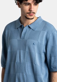 Polo de color azul claro con un patrón de cuadros texturizado, manga corta y cuello. Presenta dos botones y un pequeño logo en el pecho.