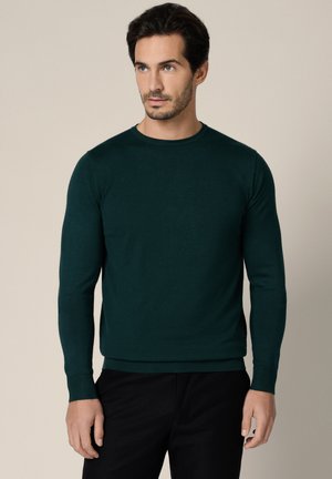 Ciemnozielony, długi sweter z dzianiny z okrągłym dekoltem i ściągaczami przy mankietach, zestawiony z czarnymi spodniami, na neutralnym tle.